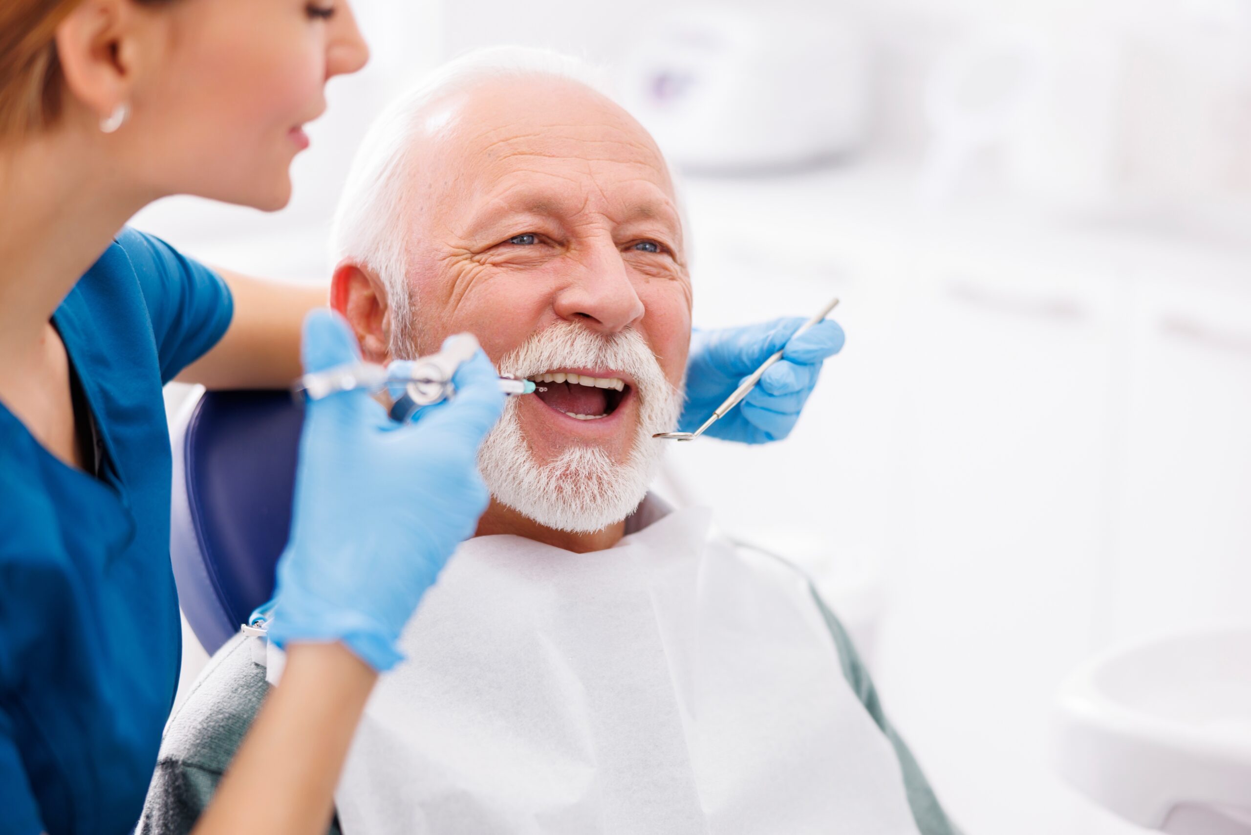 An elderly man with white hair, sat in a dentist chair with his mouth open. A female dentist is injecting his gum with a numbing solution and using a dental mirror to see the inside of his mouth.