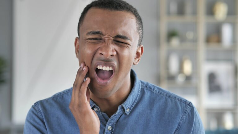 A man in a blue shirt touching his jaw with his right hand, wincing in pain due to toothache