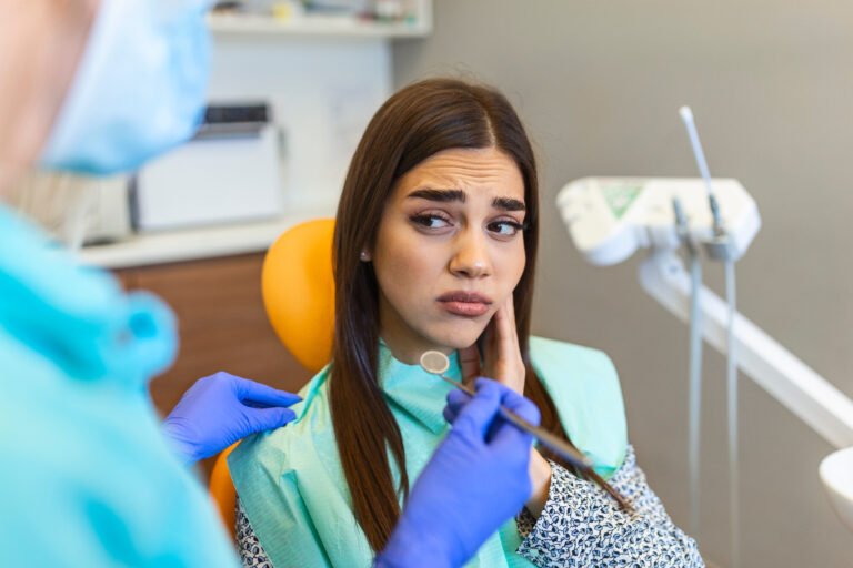 A young woman in a medical gown touching her jaw with her left hand indicating pain from a failed root canal