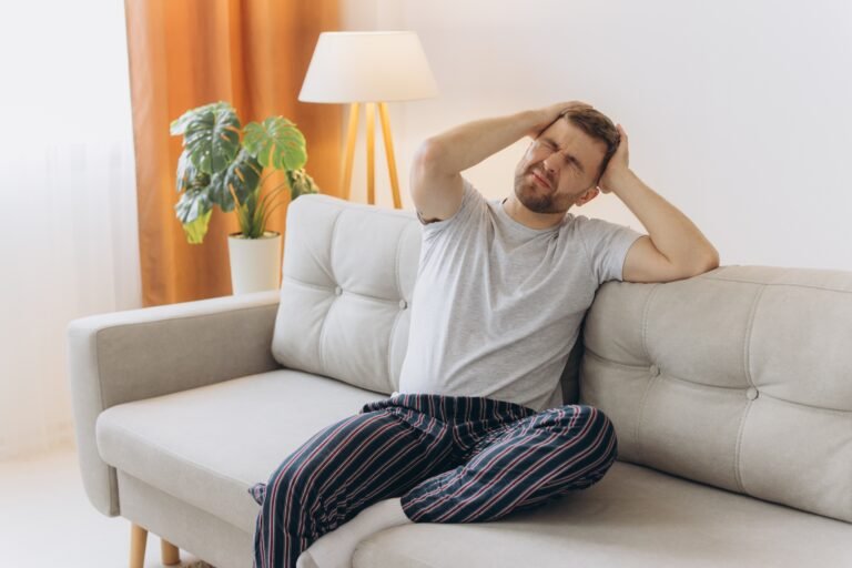 A young man sat on a sofa holding his hands to his head and wincing in pain from a headache.